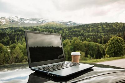 Laptop and coffee cup on a vehicle rooftop with forested mountains in the background